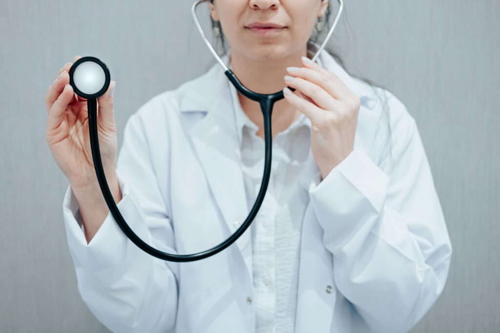 Close-up of a medical professional wearing a white lab coat and holding a stethoscope, with one hand lifting the chest piece toward the camera while the other adjusts the earpieces, set against a neutral light gray background.
