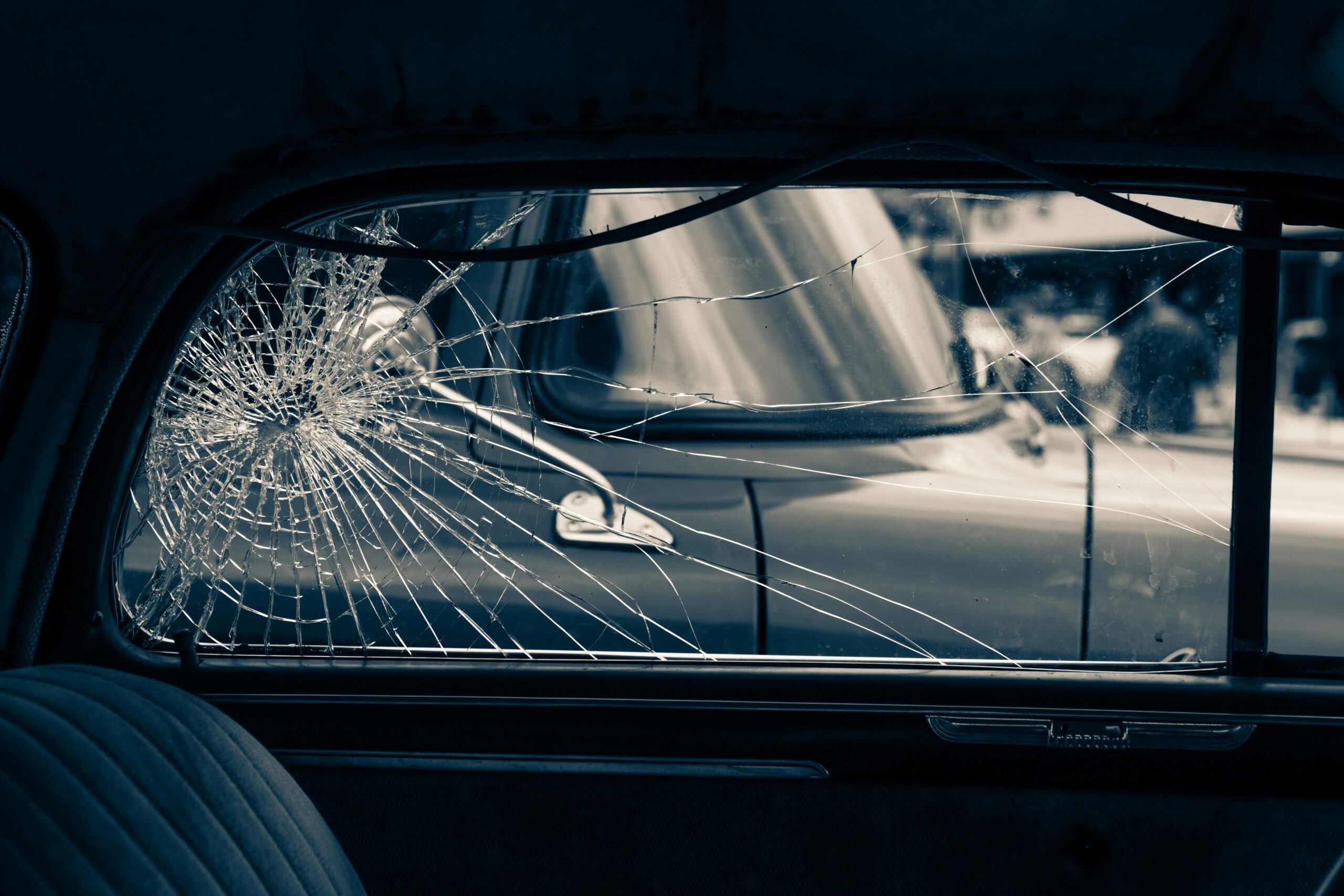 Cracked rear passenger-side car window viewed from inside the vehicle, with spiderweb fractures spreading across the glass and an intact vintage car visible outside through the damaged window, creating a muted, somber scene that suggests the aftermath of a collision.