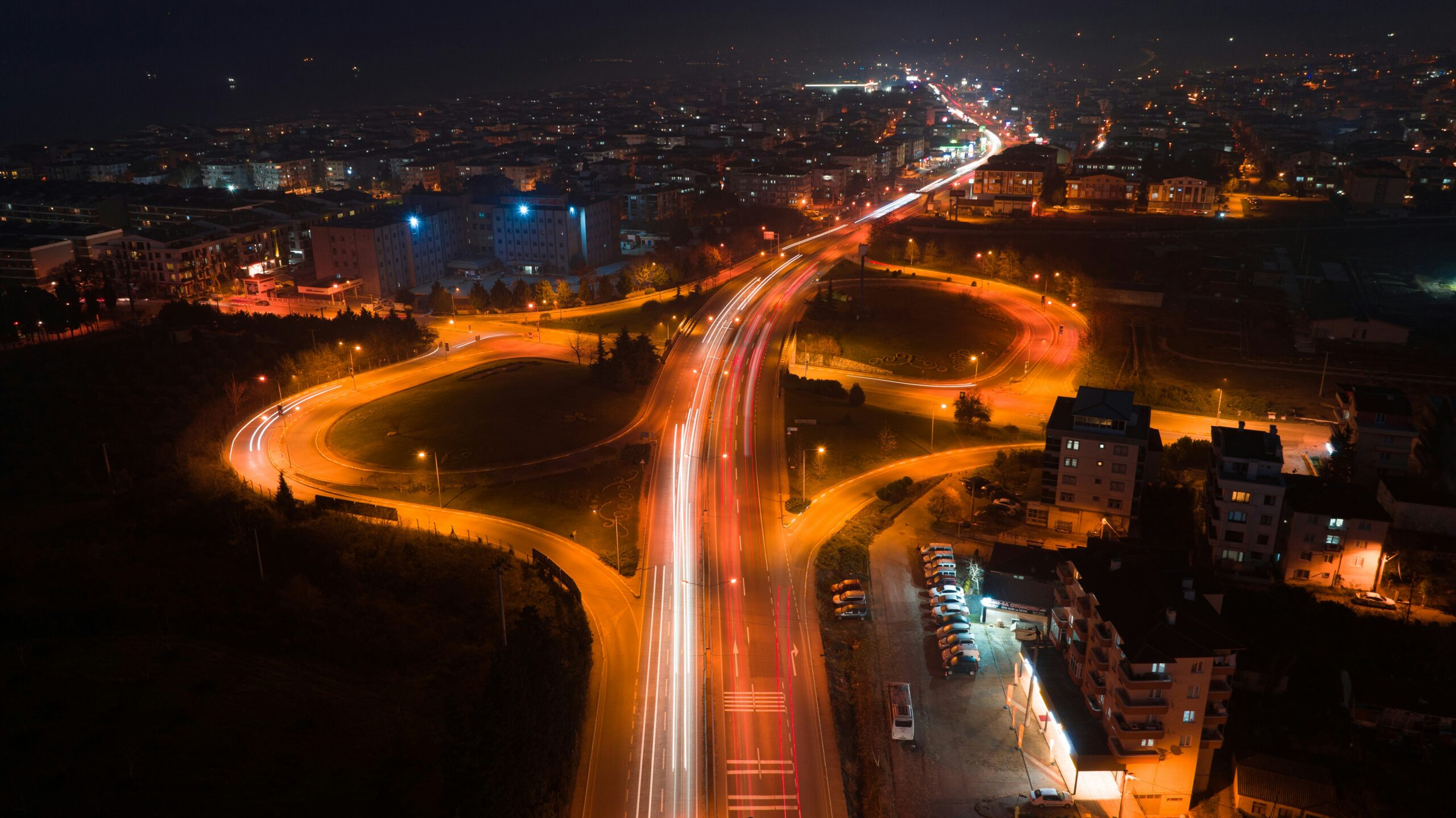 Aerial nighttime view of a sprawling city roadway with multiple curved interchanges and looping ramps, long-exposure light trails from vehicles forming bright white and red lines along the main highway, warm orange streetlights illuminating grassy medians and road edges, clusters of apartment buildings and residential blocks glowing with scattered window lights, parking lots and side streets visible near the road, and the city stretching into the distance under a dark sky.