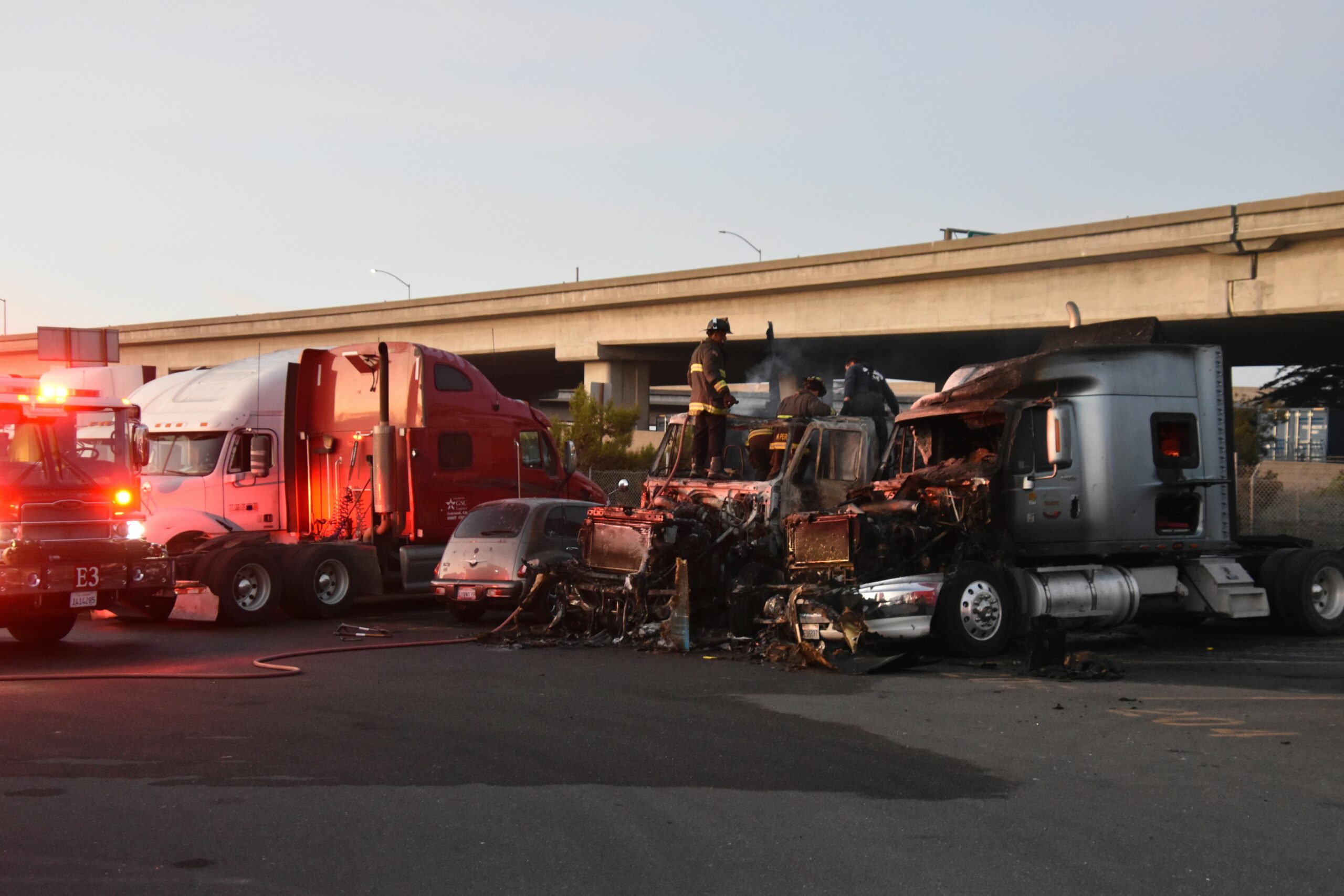 Firefighters stand atop the charred remains of multiple vehicles involved in a severe highway collision, with two heavily damaged semi-trucks and a burned passenger car stopped beneath an overpass, emergency lights from a fire engine glowing red on the roadway as smoke residue, debris, and scorch marks cover the pavement and vehicle fronts in early evening light.