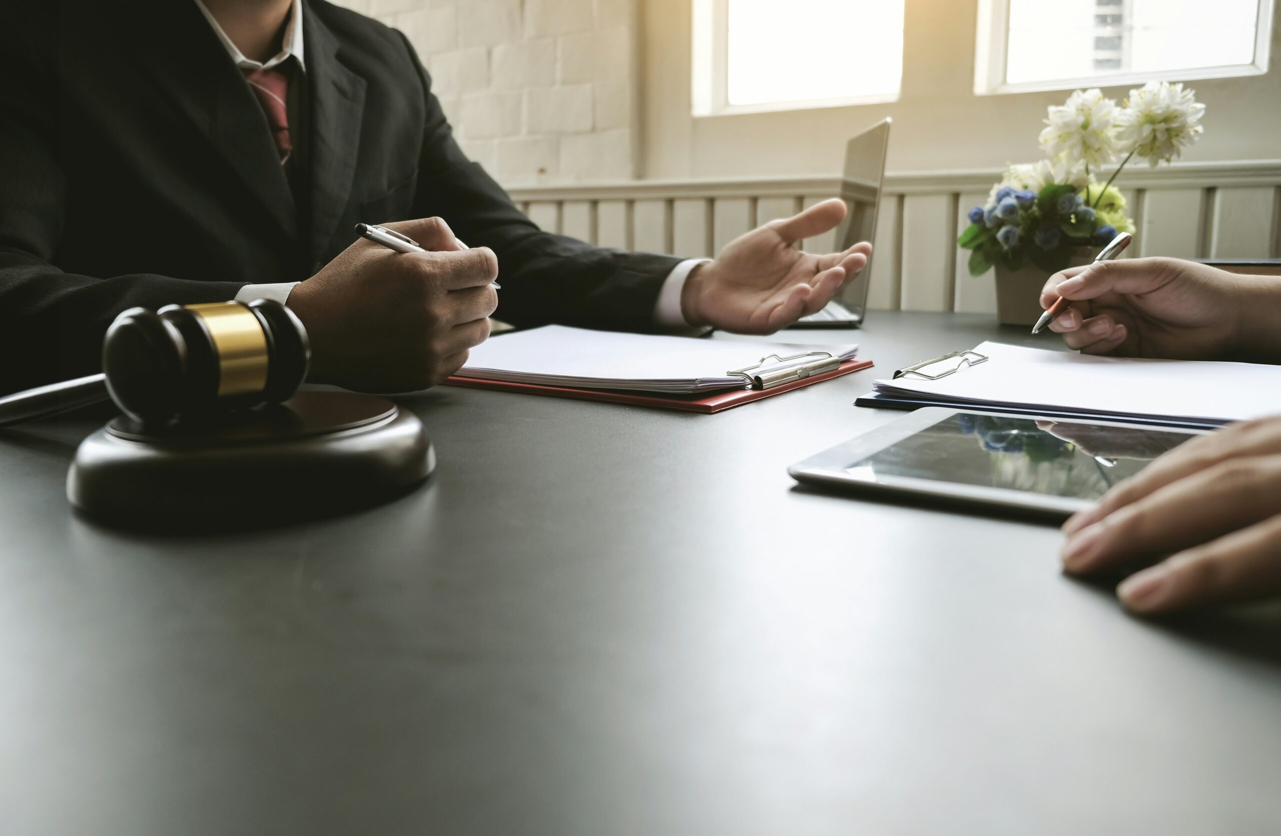 Close-up of a professional meeting at a desk where a person in a suit gestures while holding a pen, legal documents and clipboards lie on the table, a judge’s gavel sits in the foreground, and another person takes notes nearby, suggesting a legal consultation or case discussion.