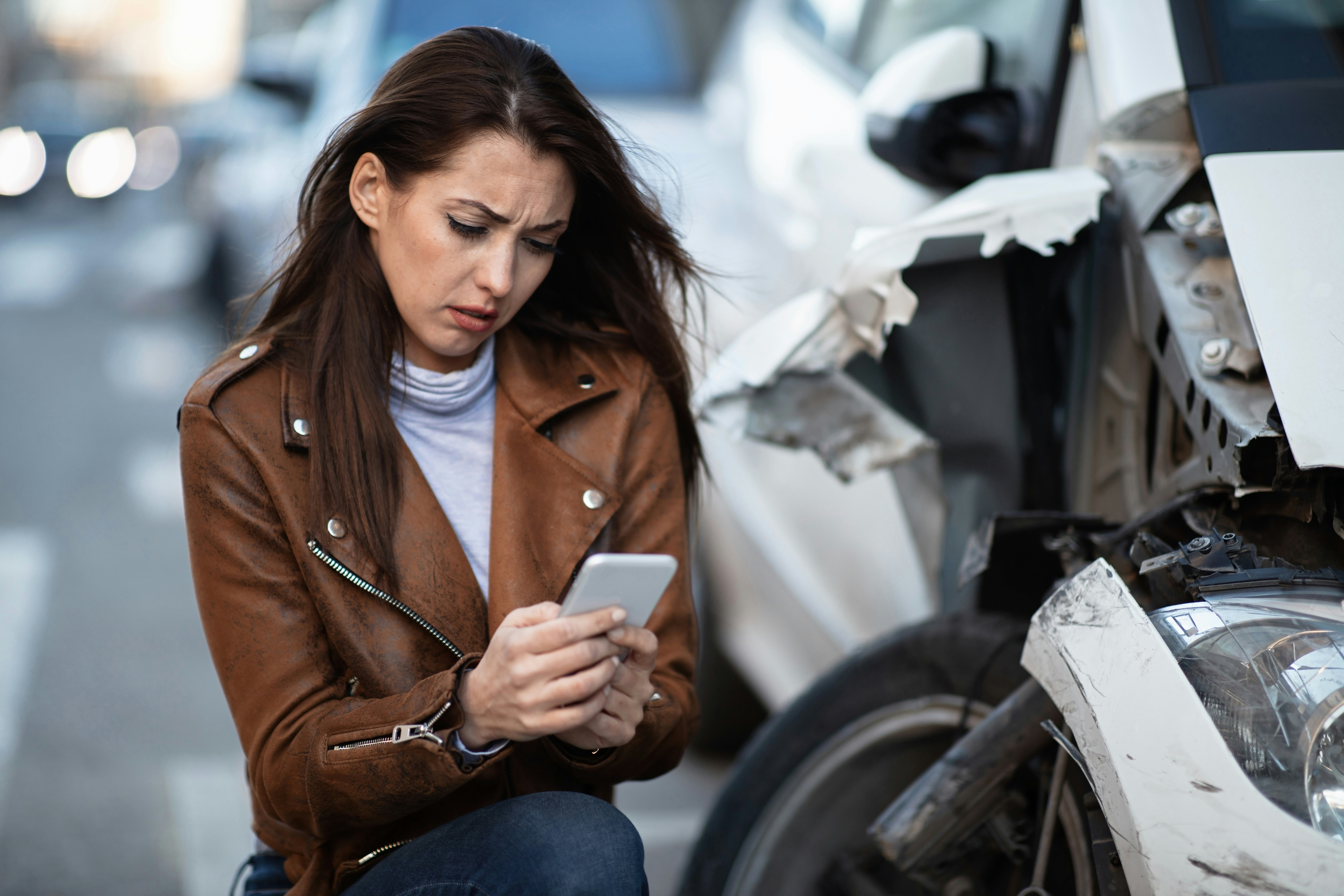 Woman crouching beside a heavily damaged white car on a city street, wearing a brown leather jacket and jeans, holding a smartphone with a worried, focused expression as she looks at the screen, with the car’s front end visibly crushed and broken, exposed internal components and torn body panels near the wheel, blurred traffic and vehicles in the background suggesting an active roadway after a recent car accident.