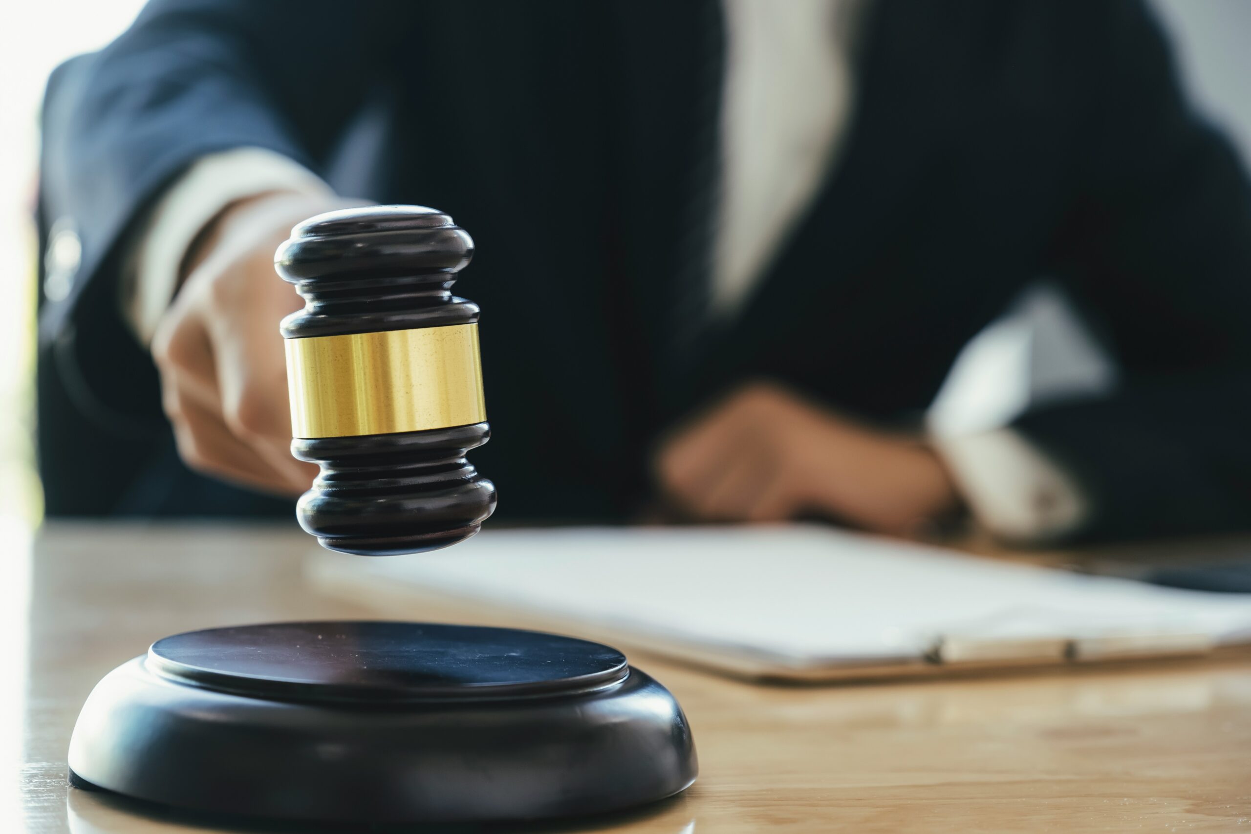 Close-up of a judge’s hand holding a wooden gavel mid-strike above a sound block on a desk, with legal documents blurred in the background, symbolizing a court ruling or legal decision.