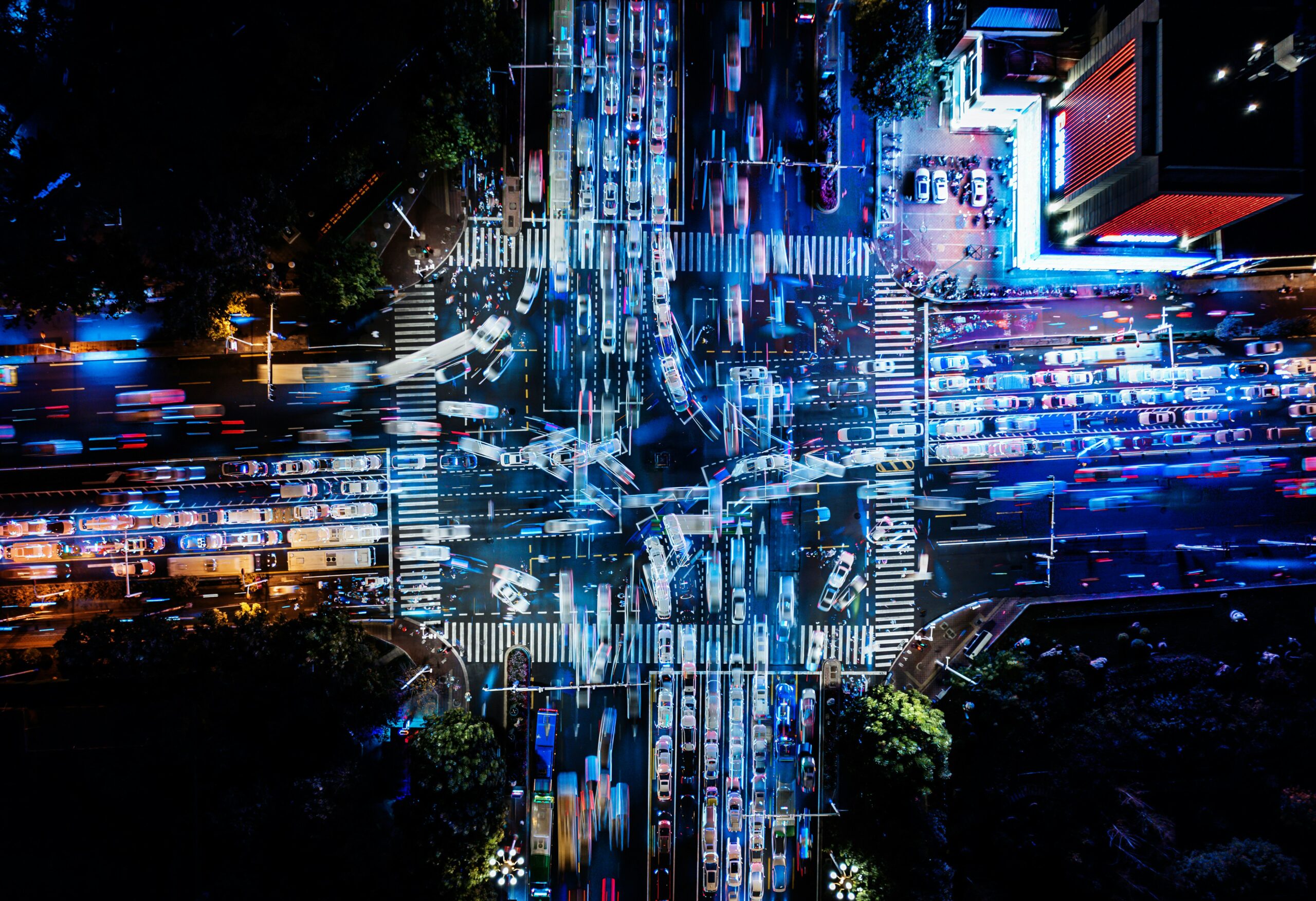 Aerial night view of a large city intersection with multiple lanes of traffic crossing in all directions, long-exposure light trails from cars forming streaks of blue, white, and red, illuminated crosswalks and traffic signals visible, dense vehicle congestion at each approach, surrounding buildings glowing with neon and streetlights, and patches of dark green trees framing the crossroads from above.