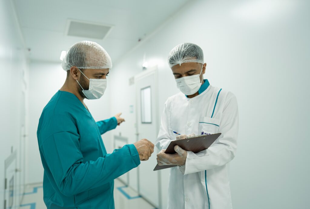 Two healthcare workers wearing surgical masks and hair caps stand in a hospital corridor, with one in scrubs pointing down the hallway while the other in a white coat writes notes on a clipboard, suggesting coordination or discussion of medical tasks.