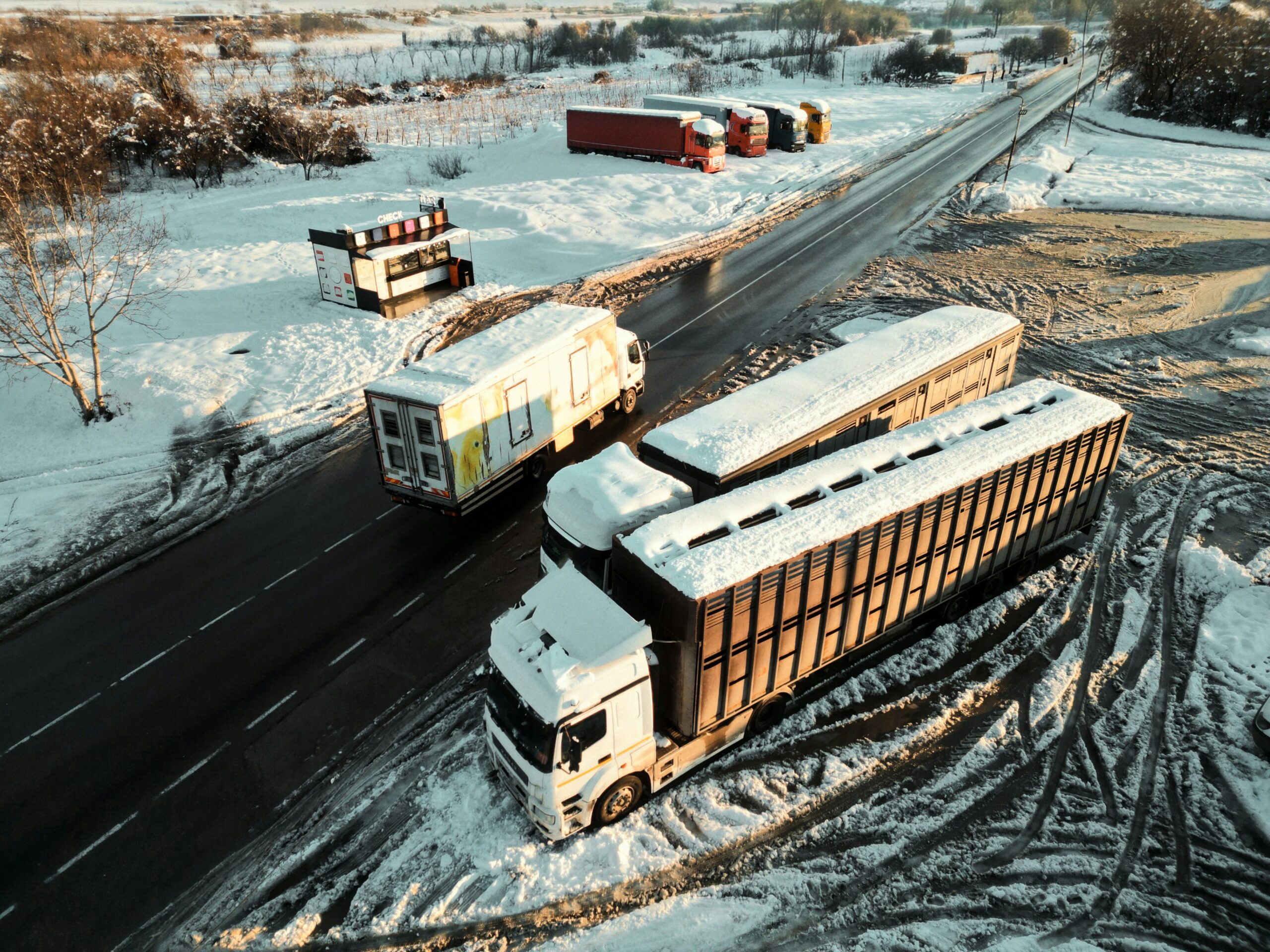 Aerial view of several large cargo trucks traveling and parked along a snow-covered rural roadway, with tire tracks etched into icy ground, trailers coated with snow, and a small roadside structure nearby, surrounded by winter fields, bare trees, and patches of frozen terrain under low, warm sunlight.